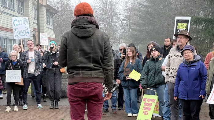 Une femme de dos avec une tuque parle à une foule. Plusieurs ont des affiches contre une hausse des droits de scolarité et la réduction des services. 