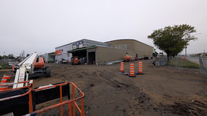 Images extérieures des travaux du nouvel aréna avec de la machinerie et des cônes au premier plan.