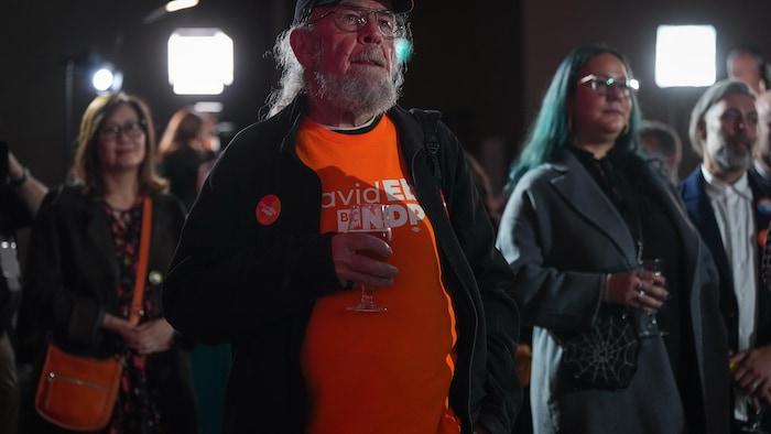 Un homme en t-shirt orange écoute un discours en tenant un verre.