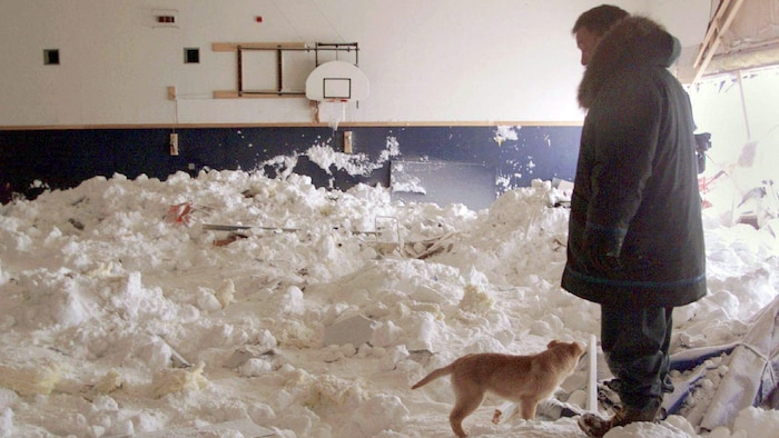 Un homme et un chien se tiennent dans la neige et constatent les dégâts causés par une avalanche qui a détruit le gymnase de l'école de Kangiqsualujjuaq, au Nunavik.