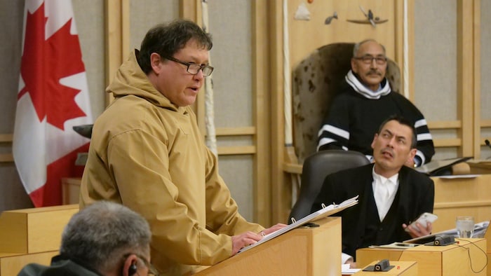 George Hickes debout à l'Assemblée législative entouré de ses collègues.