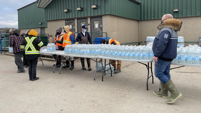 Des gens viennent chercher des caisses de bouteilles d'eau disposées sur des tables à l'extérieur d'un édifice.