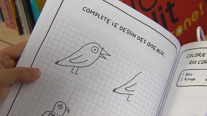Une personne tient ouvert le cahier d'activités "Nutshimit", illustrant des dessins simples de style bande dessinée pour les enfants.