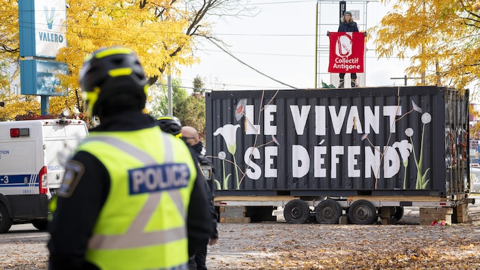 Une personne tient une bannière du collectif Antigone sur le toit d'un conteneur, où l'on peut lire : «Le vivant se défend». Des policiers observent la scène de loin.
