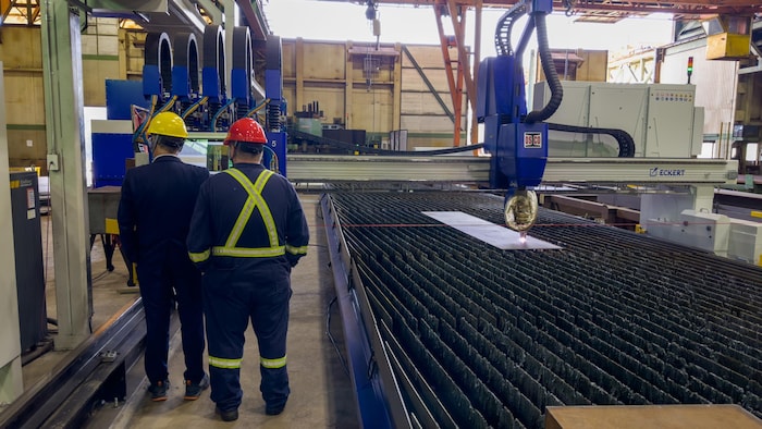 Une machine découpe une planche d'acier dans une usine. Deux hommes avec des casques de construction sont à côté de la machine.