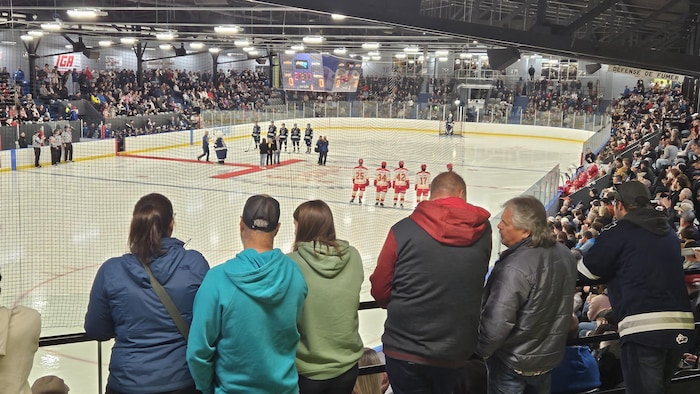 Un aréna rempli de spctateurs assis et debours devant la glace où sont alignés les joueurs des deux équipes de hockey pendant que des dignitaires sont sur le tapis rouge avant le match. 