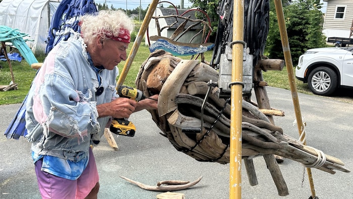 A man uses a drill to attach a piece of beach wood to a giant sculpture in a back of he sculpts giant birds beach new the house. he sculpts giant birds beach