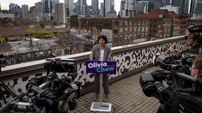 Olivia Chow, sur une terrasse devant un secteur de la ville de Toronto à l'arrière-plan, donne un discours devant de nombreuses caméras.