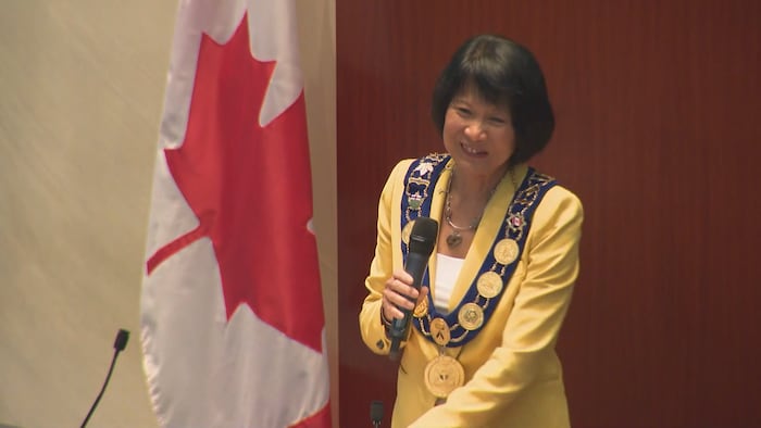 عمدة تورونتو الجديدة أوليفيا تشاو تلقي كلمة حاملة ميكروفوناً وإلى جانبها علم كندا.