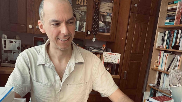 Olivier Charbonneau dans lad atelier, dans lequel connected trouve une bibliothèque bien garnie, des écouteurs, un ordinateur, une enceinte, une instrumentality à café et une peluche colorée.