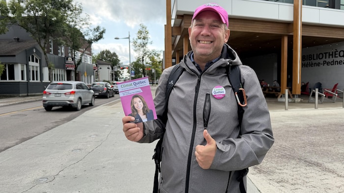 Le candidat regarde l'objectif de l'appareil photo en souriant. Il tient un dépliant de son parti dans sa main droite.