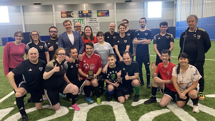Une photo de groupe rassemble une vingtaine de personnes sur un terrain de soccer intérieur. Plusieurs portent des uniformes de sport, dont un homme au centre qui tient une plaque des Special Olympics Unified Sports. L'ambiance est souriante et conviviale.
