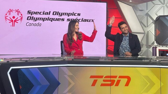 Deux animateurs, une femme en rouge et un homme en veston, saluent de la main sur un plateau de télévision de T S N. Derrière eux, un écran affiche le logo d'Olympiques spéciaux Canada sous les éclairages du studio.