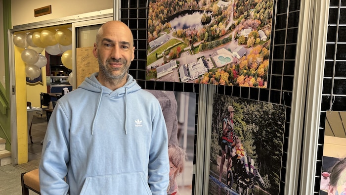 Olivier Piquer pose devant un paravent sur lequel sont accrochées des photos dont une du terrain de Cité-Joie vu des airs. Derrière lui, des ballons bordent le cadre d'une porte menant à une autre salle.