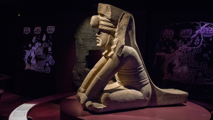 Statue en pierre représentant une personne assise par terre, bras tendus devant ses genoux. 