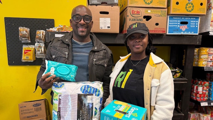 Omar Smith et une femme se tiennent dans une épicerie caribéenne devant des étagères remplies de produits alimentaires. Ils tiennent des couches, des lingettes, de la literie et d'autres articles récoltés lors d'une collecte de dons.