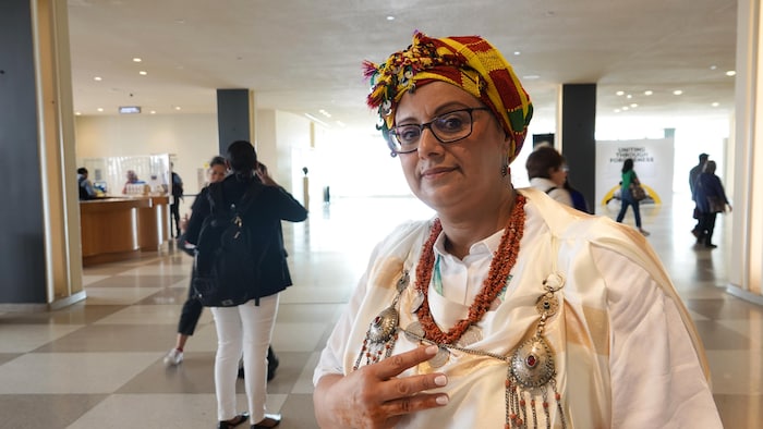 Amina Amharech pose dans une pièce de l'ONU avec un foulard traditionnel coloré sur la tête et des bijoux.