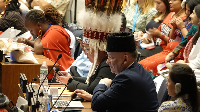 Eskender Bariiev porte un chapeau traditionnel et est assis devant un micro, entouré par de nombreux Autochtones.