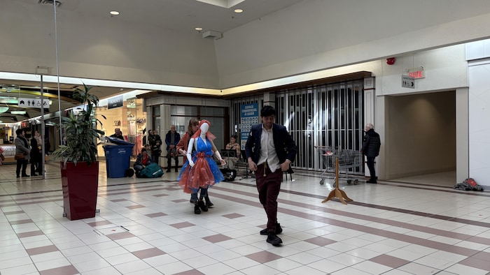 Dans l'allée d'un centre commercial, un homme marche au premier plan devant deux personnes transportant un mannequin vêtu de plastique bleu et rouge. Quelques spectateurs observent la scène près de boutiques fermées et d'un bac de recyclage bleu.