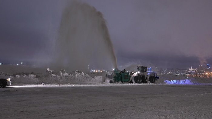 Dans la nuit, avec les lumières du centre-ville de Québec au loin, une souffleuse dégage un stationnement en soufflant la neige dans un grand jet en hauteur. 