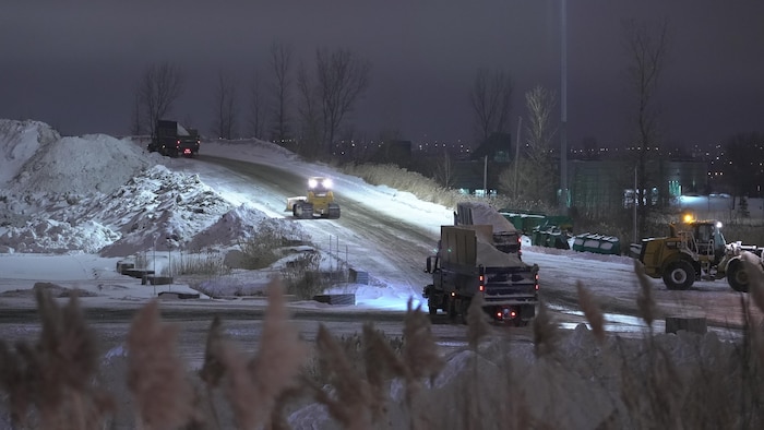 Dans la noirceur, des camions remplis de neige avancent les uns derrière les autres vers le haut d'une colline où ils déchargent la neige. 