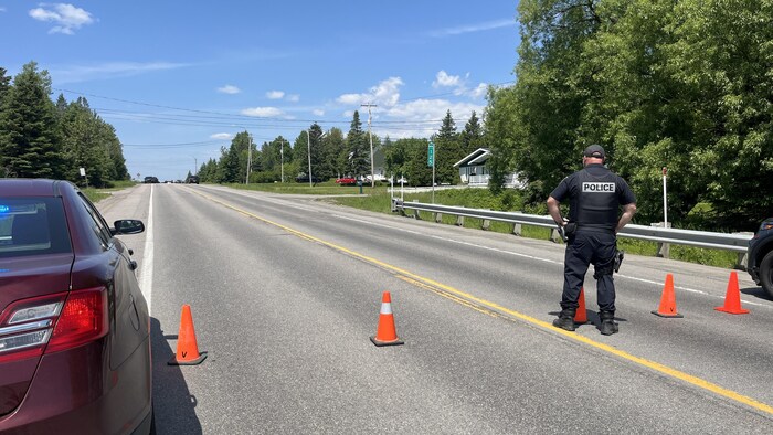 Un policier de dos se tient debout au milieu d'une route bloquée par des cônes oranges.