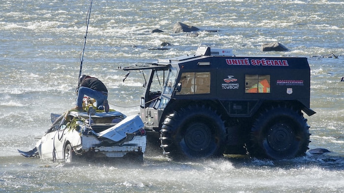 Un homme attache des câbles au toit de la voiture à côté de laquelle se trouve le camion amphibie.