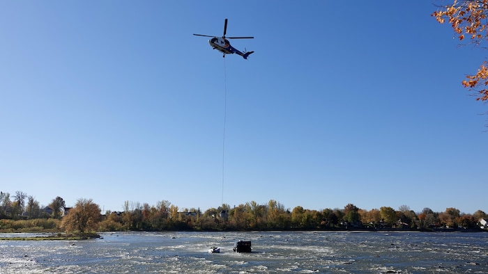 Un long câble relie la voiture à un hélicoptère qui s'apprête à la sortir de la rivière.