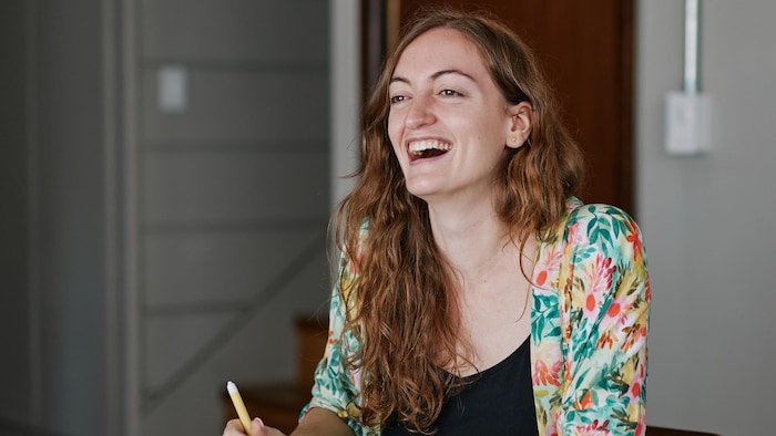 Ophélie Surcouf est assise avec un crayon à sa main et un cahier sur la table devant elle. Elle sourit.
