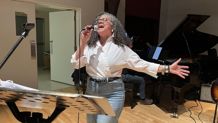 La chanteuse tient un micro et chante devant un pianiste. Elle est sur scène.