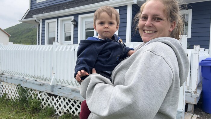 Une femme tient un enfant dans ses bras devant une maison.
