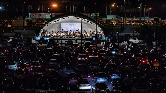Des voitures sont devant une scène sur laquelle jouent des musiciens et des musiciennes. 