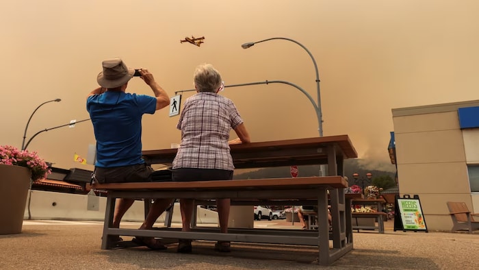 Un couple assis à une table observe un bombardier d'eau sous un ciel enfumé.