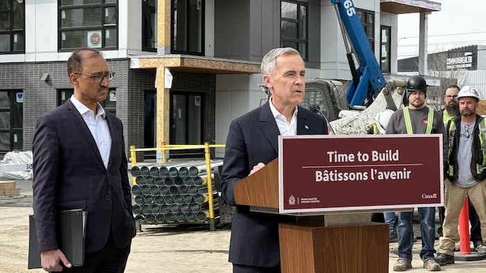 Mark Carney parle à un podium à l'extérieur. Le maire d'Edmonton Amarjeet Sohi se tient derrière lui. 