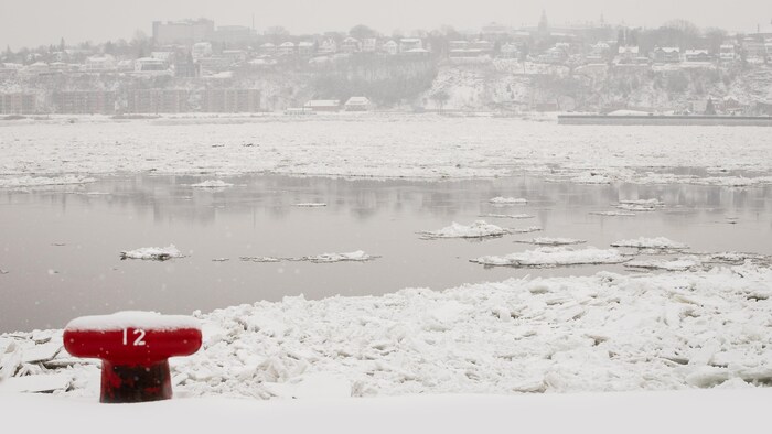 Une ancre rouge en bordure du fleuve. En arrière-plan, une vue sur le fleuve plein de glaces et la ville de Lévis.