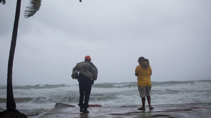 Deux hommes sont sur une place, devant une mer déchaînée.