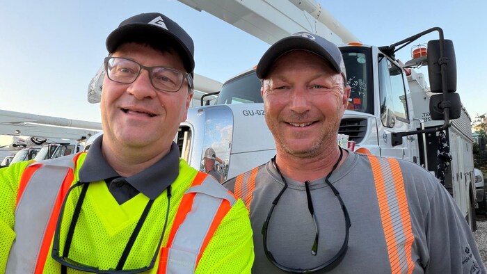 Louis Léger et Roger LeBlanc sont debout devant un camion de monteur de lignes. Ils portent tous les deux une casquette et sourient en regardant la caméra.