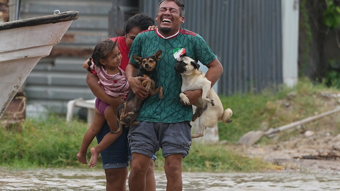 Un homme qui tient deux petits chiens collés sur lui, marche en riant dans la rue inondée, suivi par une femme portant une petite fille souriante, l'eau montant jusqu'aux mollets.