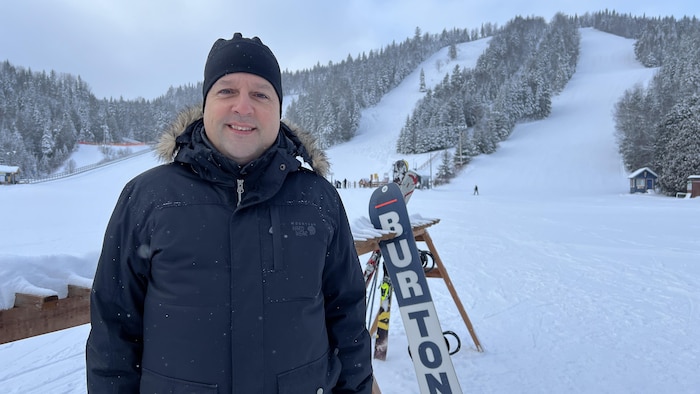 Il est devant les pentes du Mont Castor, devant une installation où sont déposés skis et planches à neige.