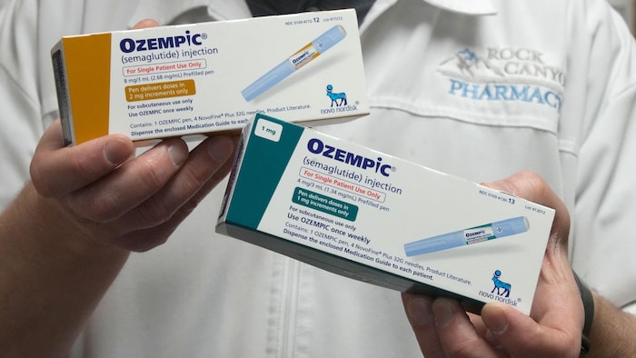 Un homme vêtu d'un sarrau de pharmacien tient dans ses mains deux boîtes d'Ozempic, qui affichent des informations sur le médicament et la photo d'un injecteur.