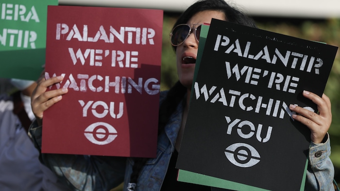 Une personne manifeste en tenant dans ses mains deux affiches disant en anglais « Palantir, nous vous surveillons ».