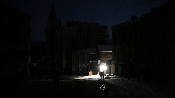 Deux piétons marchent dans une rue non éclairée, vers une lumière, au cœur de la nuit.