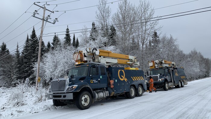 Deux camions d'Hydro-Québec sont stationnés sur le bord d'une route. Un travailleur marche entre les deux.
