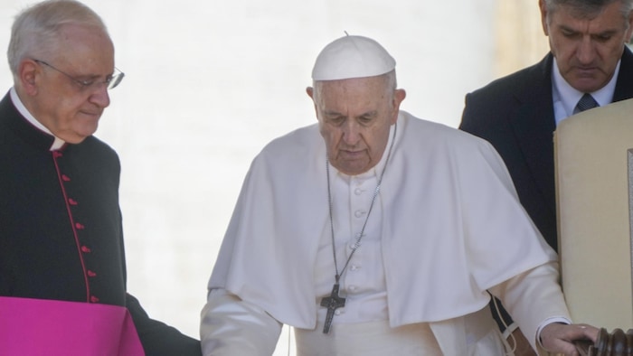 البابا فرنسيس يتكئ على عصا للمشي ويعاونه كاردينال ورجل آخر.