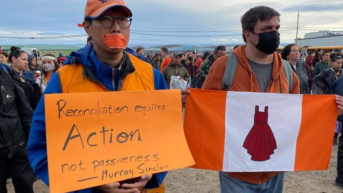 Deux personnes tiennent une pancarte et un drapeau avec des messages sur la réconciliation et une robe rouge, représentant les filles et femmes autochtones disparues et assassinées, le 29 juillet 2022 à Iqaluit.