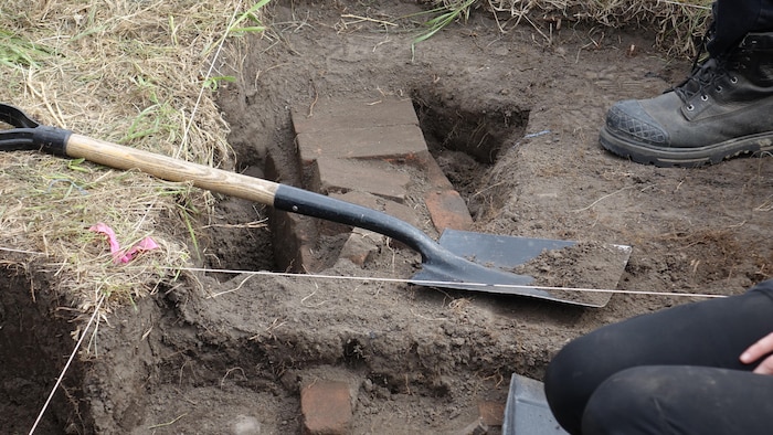 Vue en plongée d'un chantier de fouilles archéologiques où une pelle de chantier repose sur le sol en terre à côté d'une tranchée laissant apparaître de vieilles briques enfouies, tandis qu'une personne agenouillée et une botte de travail sont visibles en bordure.