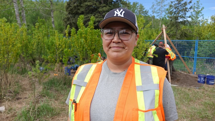 Une femme souriante posant à l'extérieur devant un chantier de fouilles archéologiques entouré d'arbres et de végétation.
