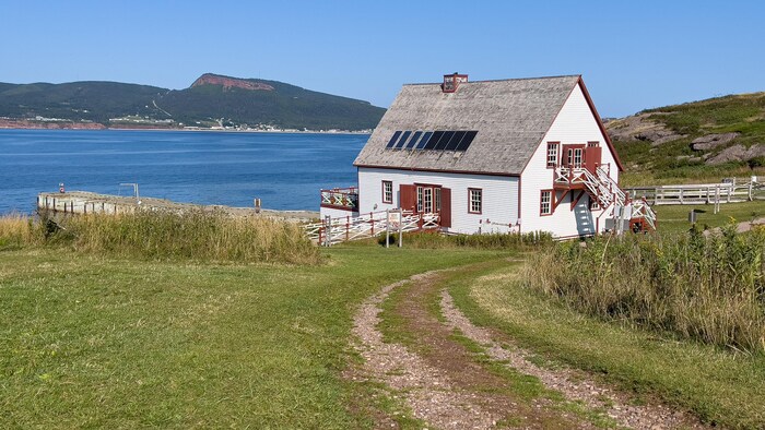 Photo prise sur l'île Bonaventure. Une maison patrimoniale se tient tout près de l'eau et d'un quai. L'autre côté de l'étendue d'eau se trouve la Ville de Percé et une montagne.