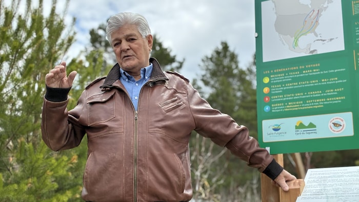Un homme aux cheveux gris, vêtu d'un manteau de cuir brun, s'exprime devant un panneau d'information sur la migration des monarques. Des conifères et un ciel clair forment l'arrière-plan de cette scène captée en extérieur.