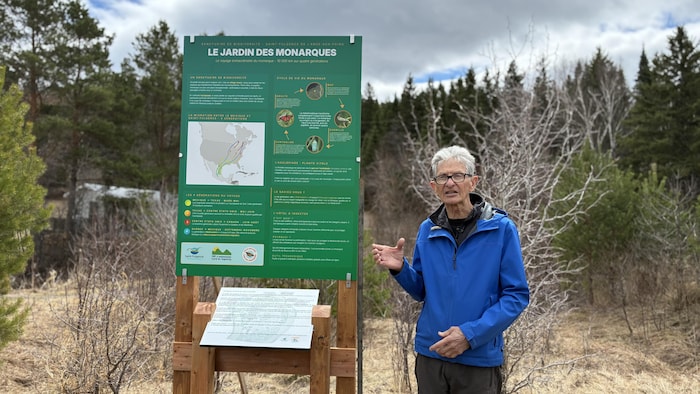 Un homme en manteau bleu gesticule à côté d'un grand panneau vert intitulé « Le Jardin des Monarques ». L'installation éducative est située à l'extérieur, entourée d'arbres et de végétation sèche sous un ciel partiellement nuageux.